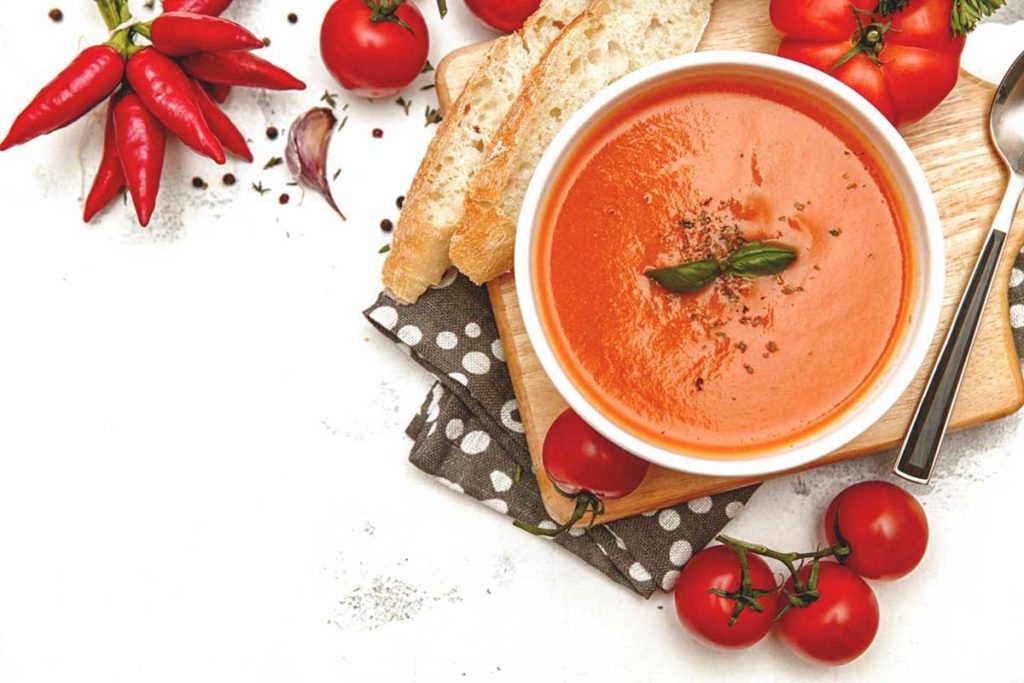 Tomato soup in a white bowl on a chopping board surrounded by tomatoes and chillies