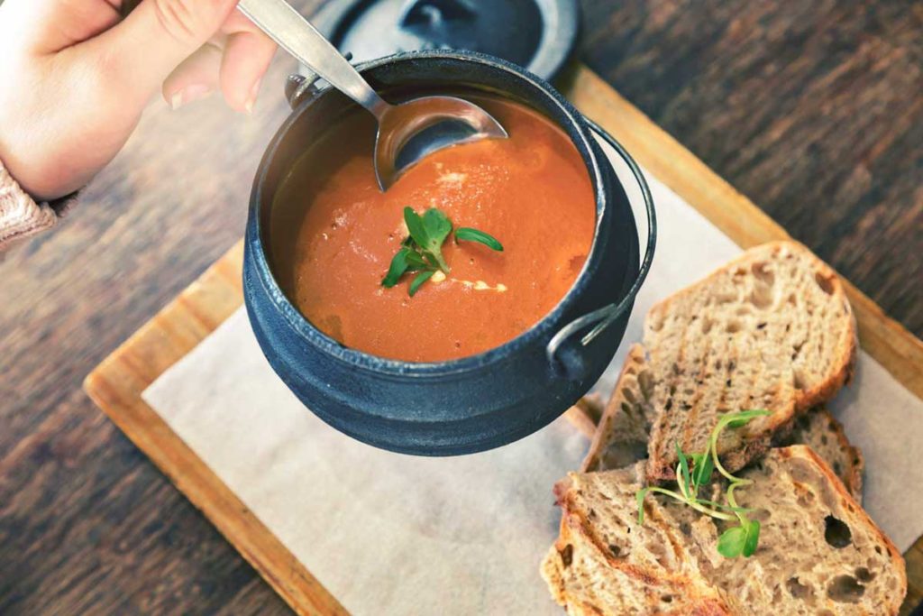 Tomato soup in a small black cauldron on a chopping board with bread