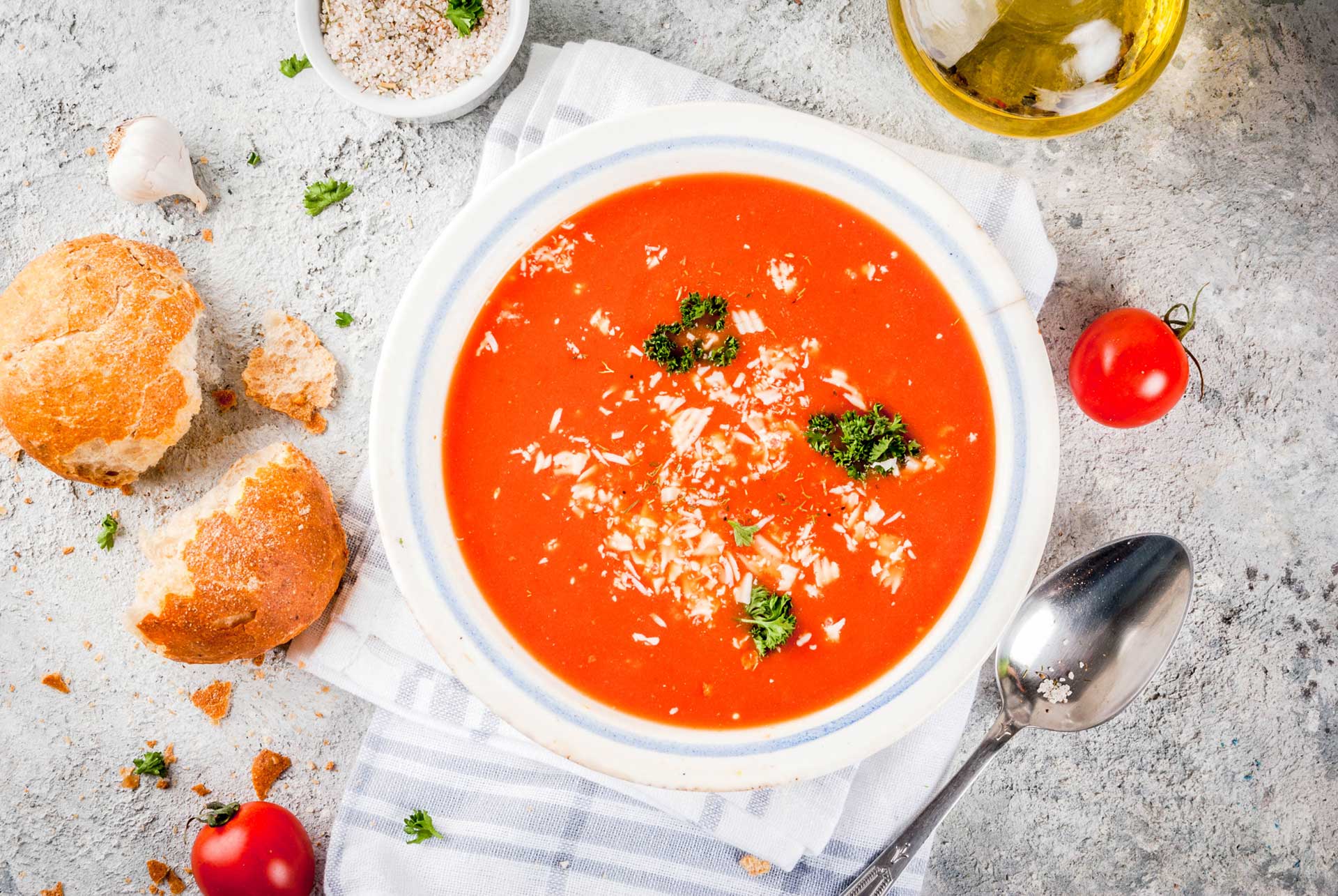 Tomato soup in a white bowl on a tea towel surrounded by tomatoes, bread and seasoning