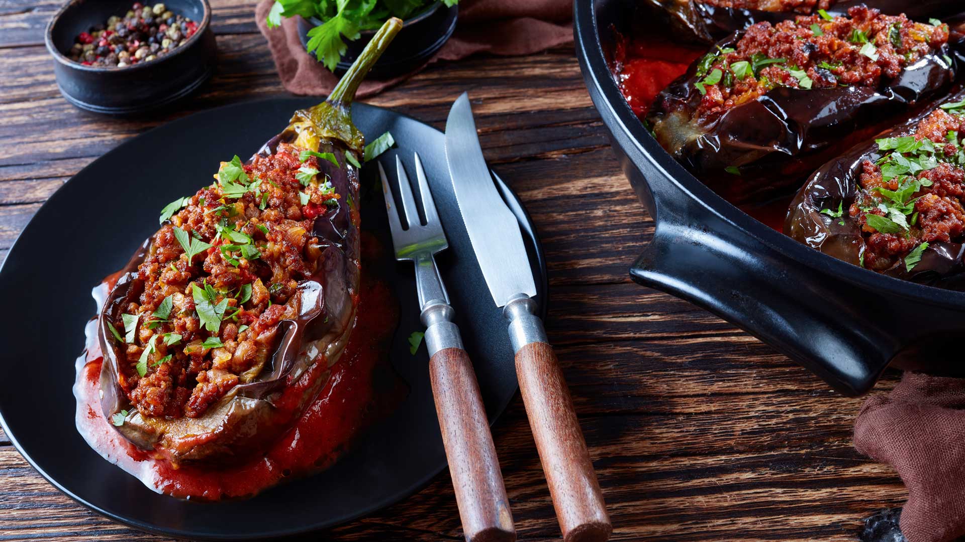 Karniyarik - Stuffed Eggplants, Aubergines with ground beef and vegetables baked with tomato sauce served on a plate with fork and knife, turkish cuisine, horizontal view from above, close-up, flatlay