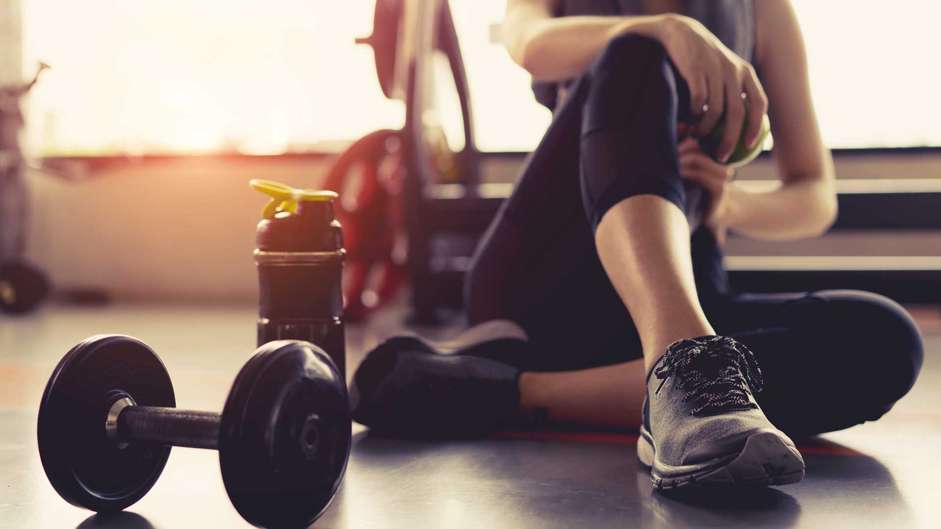 Women at gym wearing leggings and tracking shoes. She is sitting on the floor. Near her there is water bottle and dumble