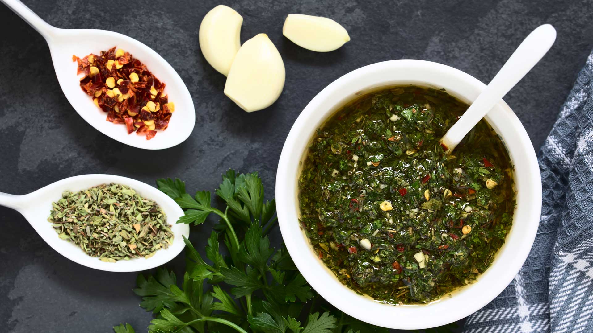 Green chimichurri sauce in a white dish on a blue cloth, surrounded by herbs, dried chillies and garlic