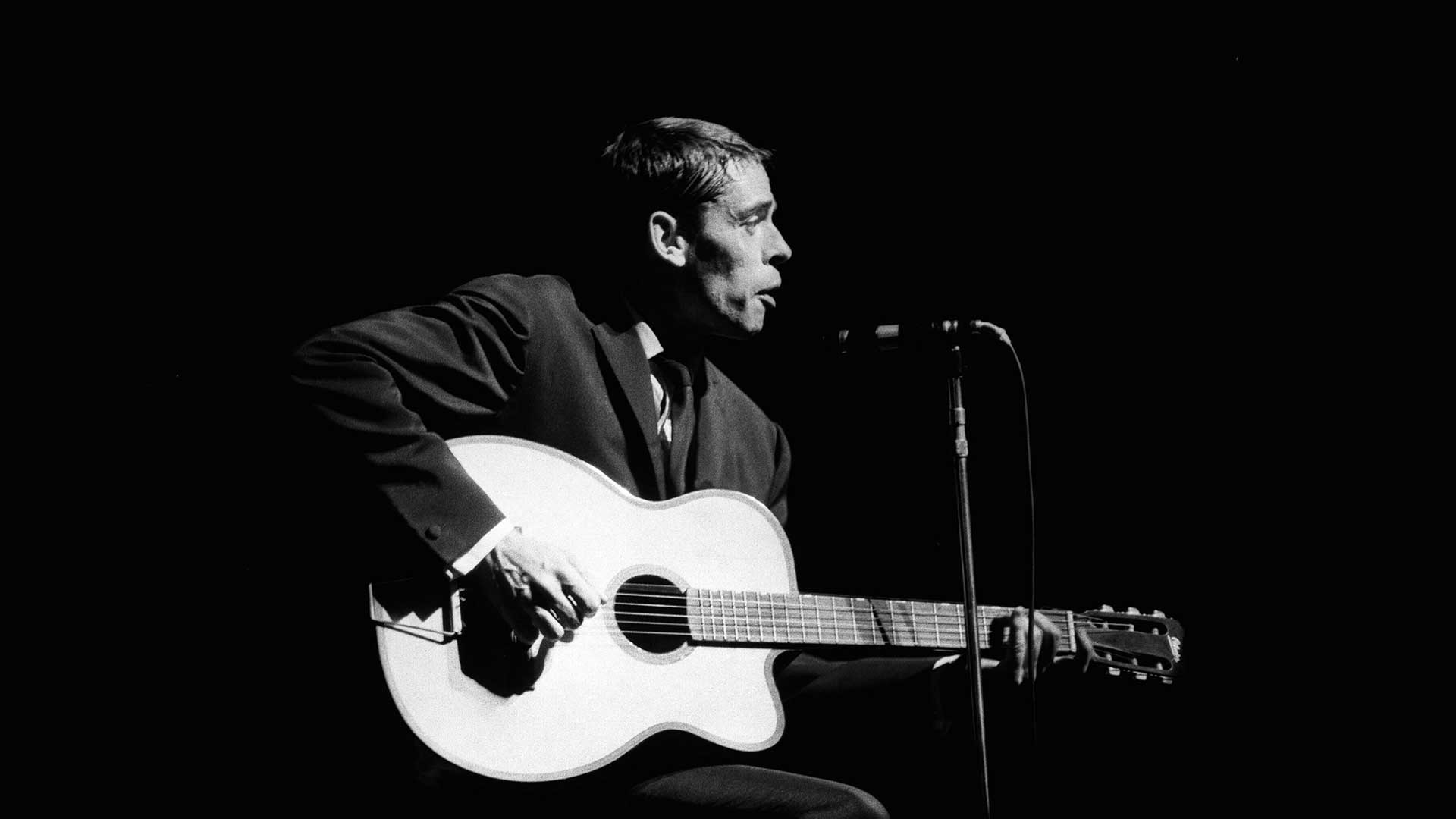 Black and white photo of Jacques Brel playing a guitar and singing into a microphone