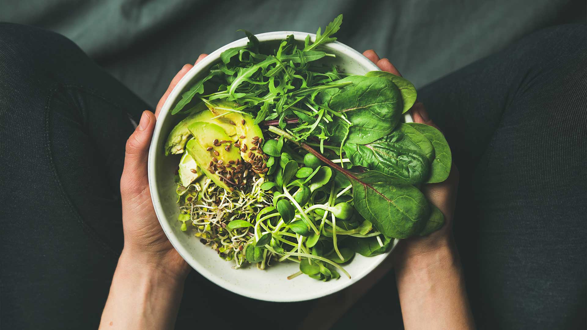 A white bowl of healthy greens and leaves held in two hands over a dark blue background