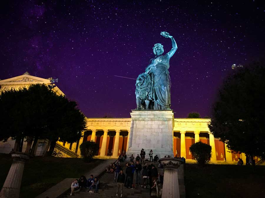 A blue/grey statue in the foreground against a background of a purple night sky with stars and an old building with columns illuminated with yellow light