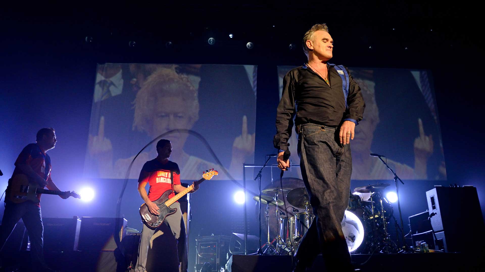 Morrisey dressed in black on stage with two backing guitarists against a blue/black background with screens showing the Queen