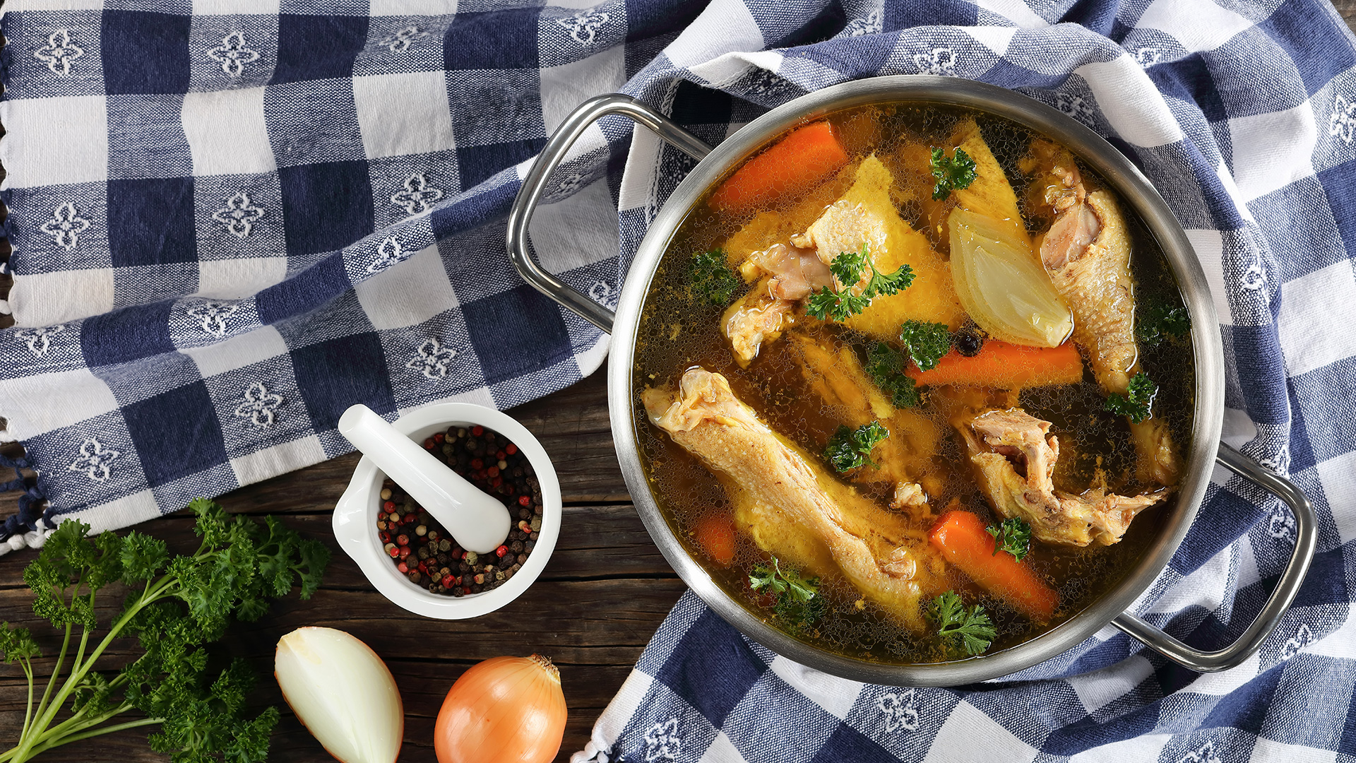 Clear Chicken broth with pieces of hen and rooster meat on bone in a metal cooking pot on wooden table with kitchen towel and ingredients on wooden table, view from above