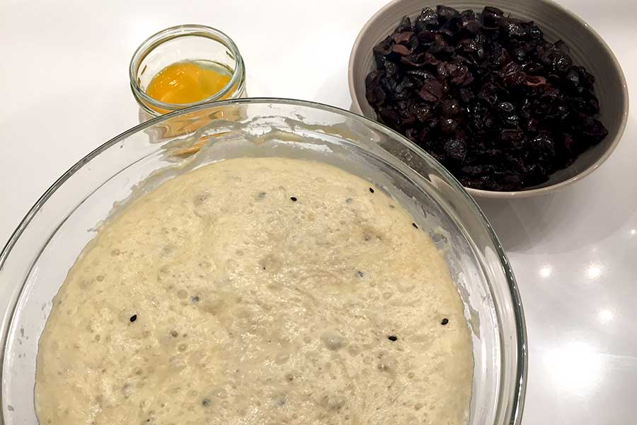 egg yolk in a bowl, dough in a bowl and black olives in a bowl on white table