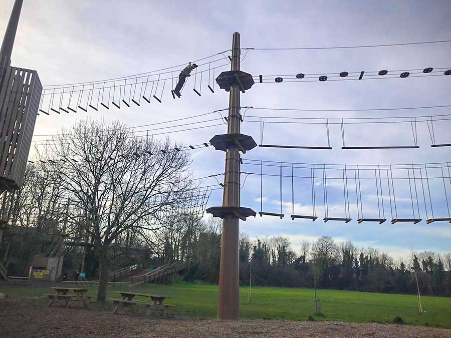 Man climbing across a high rope bridge suspended between wooden pillars against the background of a park