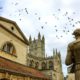 Stone statue of the roman in Antique Roman Baths complex, flying birds in sky and Abbey Cathedral at background.