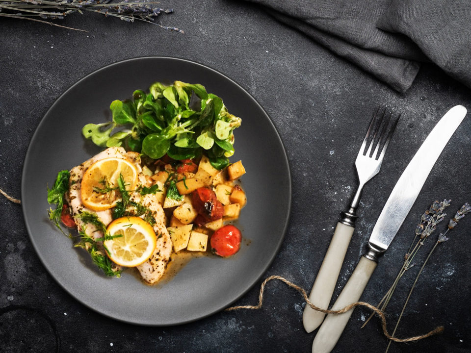 Baked Lemon & Herb Fish With Cherry Tomatoes on black plate with black dinner table and cutlery set