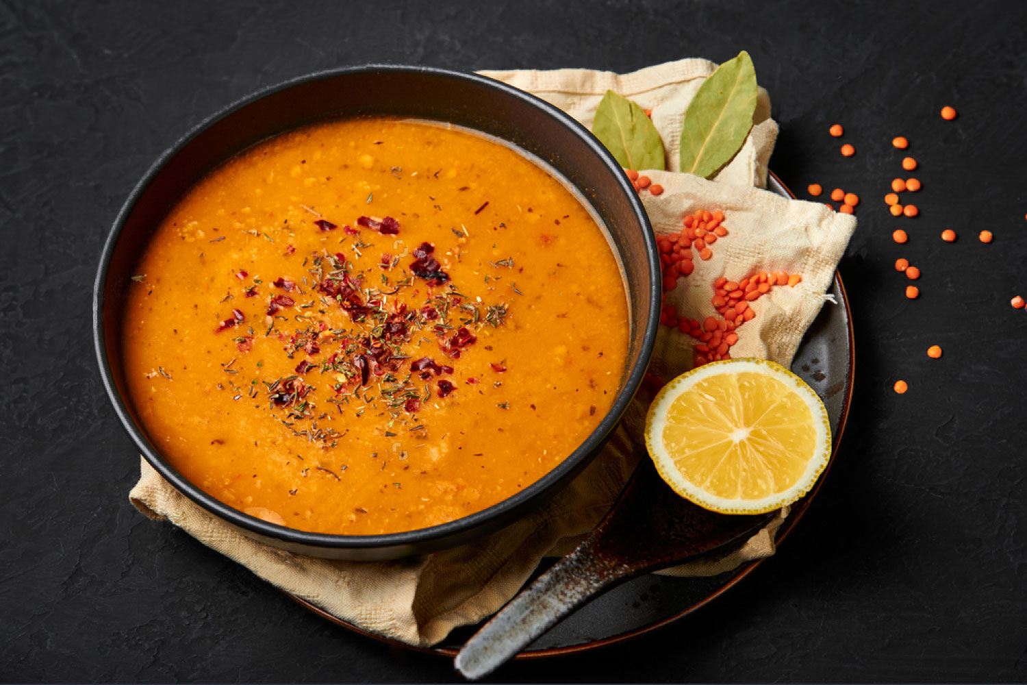 Classic Turkish Ezogelin soup in a black plate with a spoon, lemon, lentils and bayleaf. Dark background